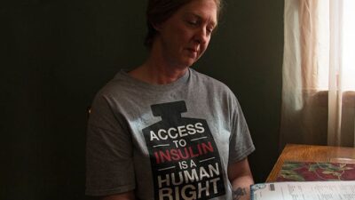 Woman sits at table looking at a paper, wearing a t-shirt that reads "Access to Insulin is a Human Right."