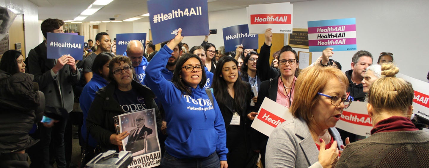 A crowd gathers around state Sen. Maria Elena Durazo before a committee hearing on legislation that would expand Medicaid coverage to adults who are in the country illegally. (Harriet Blair Rowan/California Healthline)