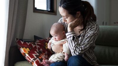 Mother sits on couch holding her newborn baby in her lap. She has a sad expression.