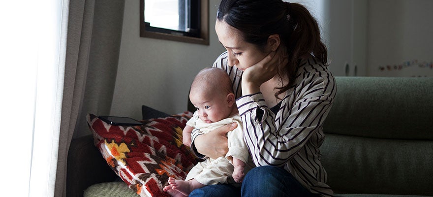 Mother sits on couch holding her newborn baby in her lap. She has a sad expression.
