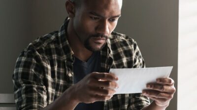 young man sitting at desk holding an envelope with a bill, looks concerned