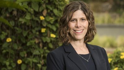 Portrait of reporter, Anna Gorman standing outdoors, against a green hedge with yellow flowers.