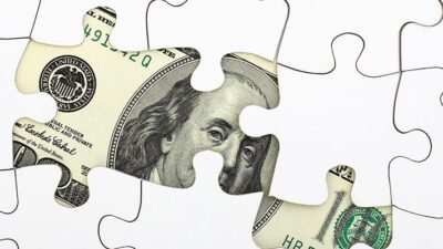 Benjamin Franklin's portrair on a one hundred dollar bill peeks out from underneath a puzzle