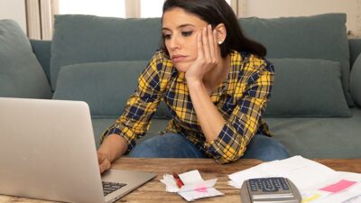 A young woman sits on a couch working at a computer, and examining her bills. She looks distressed.