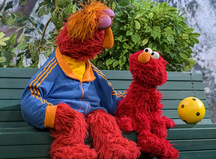 Sesame Street character Elmo sits with his father on a park bench.