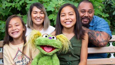 Sesame Street character, Karli, and family sit on bench.