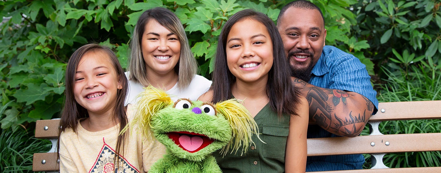 Sesame Street character, Karli, and family sit on bench.