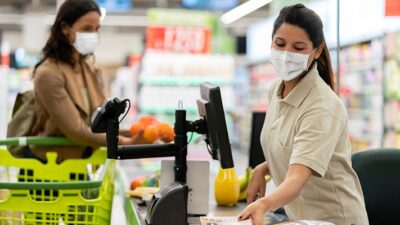 Grocery store worker wearing a surgical mask scans items at check stand as customer waits.