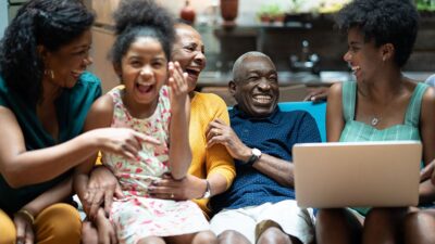 Black family sitting on couch together, looking at something on a laptop, laughing