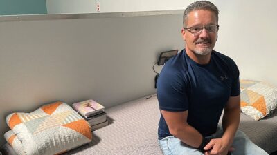 Smiling man sitting on neatly made bed with folded quilt and stack books nearby