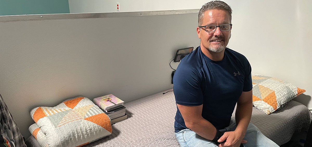 Smiling man sitting on neatly made bed with folded quilt and stack books nearby