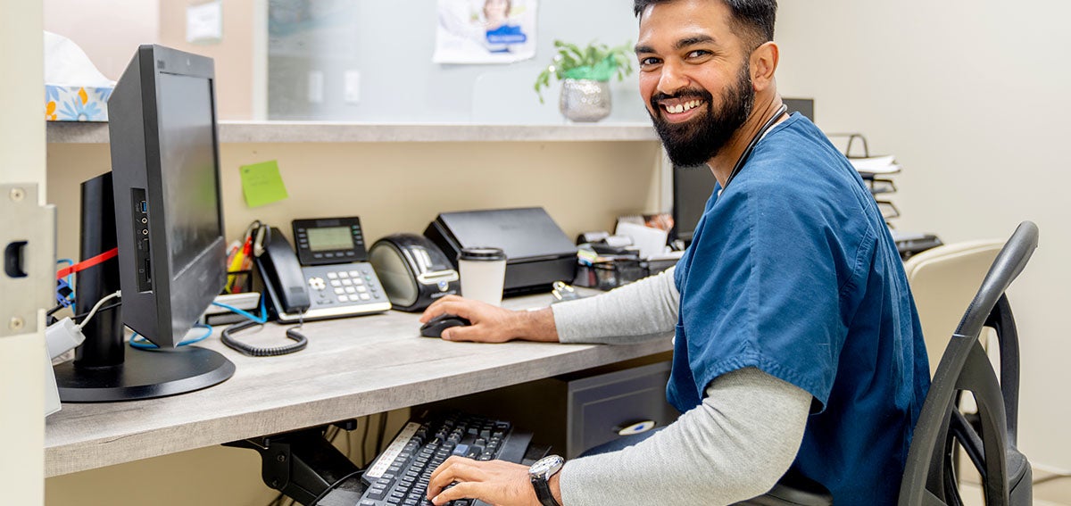 Smiling man wearing medical scrubs, working at his computer in a clinic.