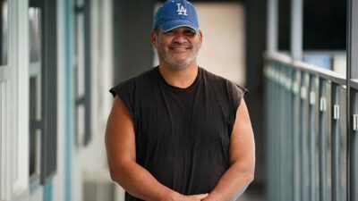 Man in baseball cap stands smiling on balcony of apartment building.