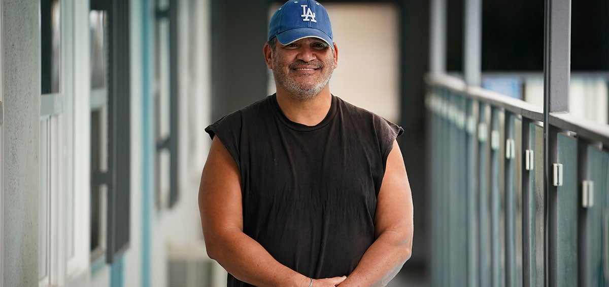 Man in baseball cap stands smiling on balcony of apartment building.