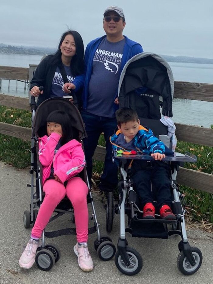 Mother and father standing near a body of water, smiling, with two children in strollers.