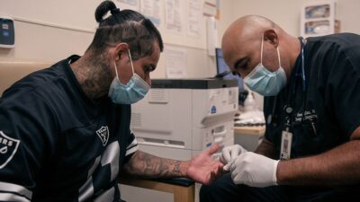 A man removes a pair of stitches from a patient's hand in a hospital setting.