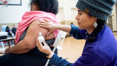 Photo of health worker attending to a patient's arm implant in a school gymnasium.