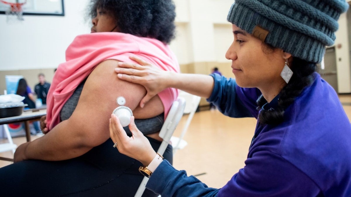 Photo of health worker attending to a patient's arm implant in a school gymnasium.