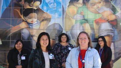 The team at Salud Para La Gente stands in front of a tile mural of motherhood.