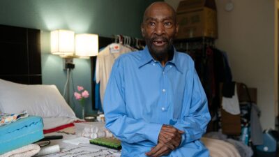Elderly man sitting on hotel bed surrounded by his belongings.