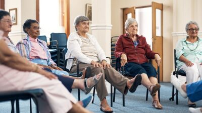 Group of senior women in assisted living facility participate in a seated exercise class.