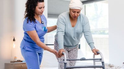 Medical practitioner helps patient walk with assistive device.