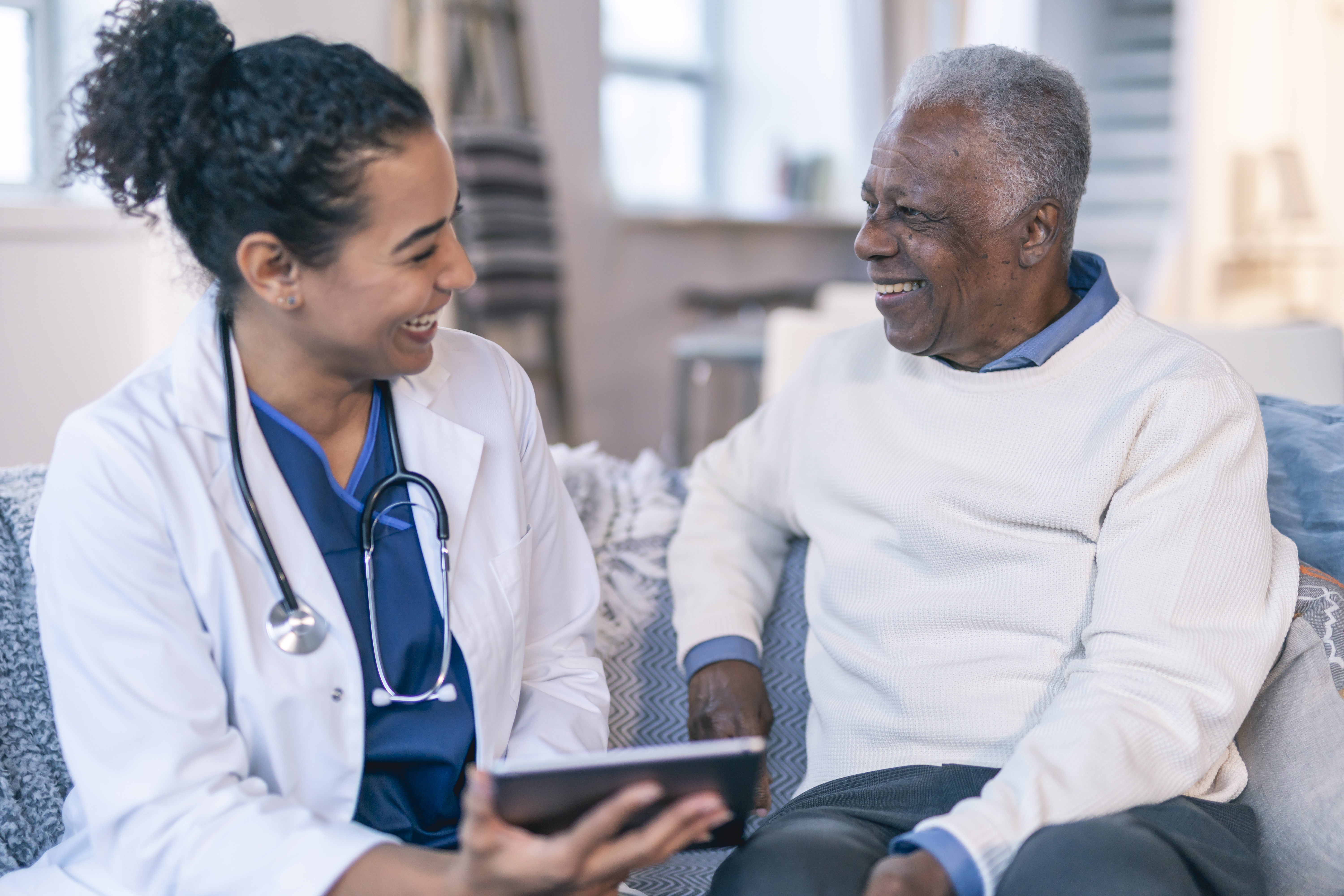 A female physician is meeting with a patient in the patient's home. The patient is a black senior adult man. The doctor and patient are seated next to each other on a couch. The doctor is holding a wireless tablet computer. The two individuals are smiling and looking at each other.