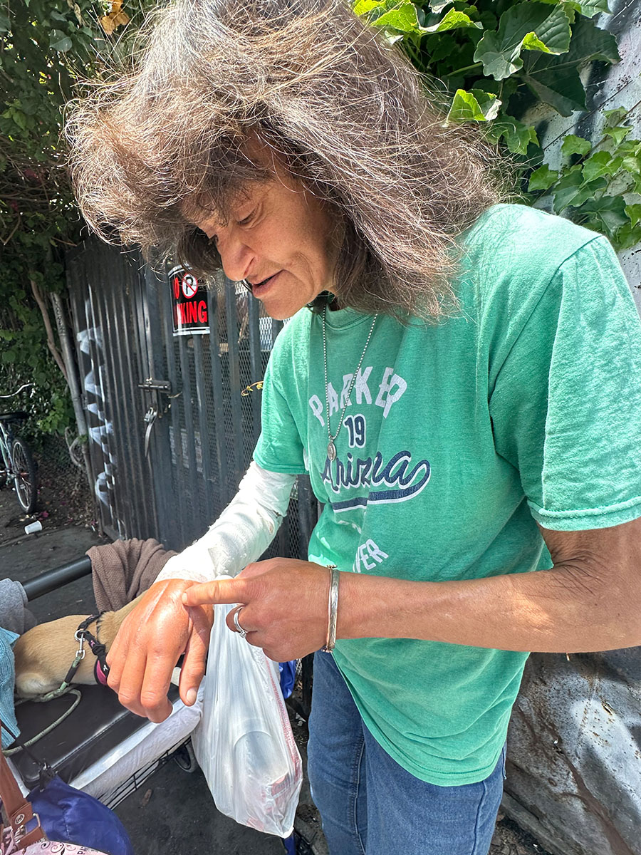 Skid Row Care Campus - A woman experiencing homelessness stands in an alley, she points to her hand which is recovering from a recent surgery.