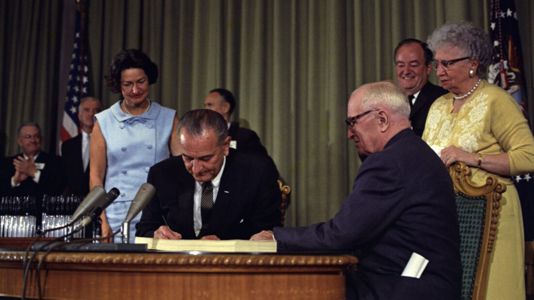 60 years of medicaid - President Lyndon B. Johnson signing the Medicare Bill at the Harry S. Truman Library in Independence, Missouri. Former President Harry S. Truman is seated at the table with President Johnson. Lady Bird Johnson stands behind the president.