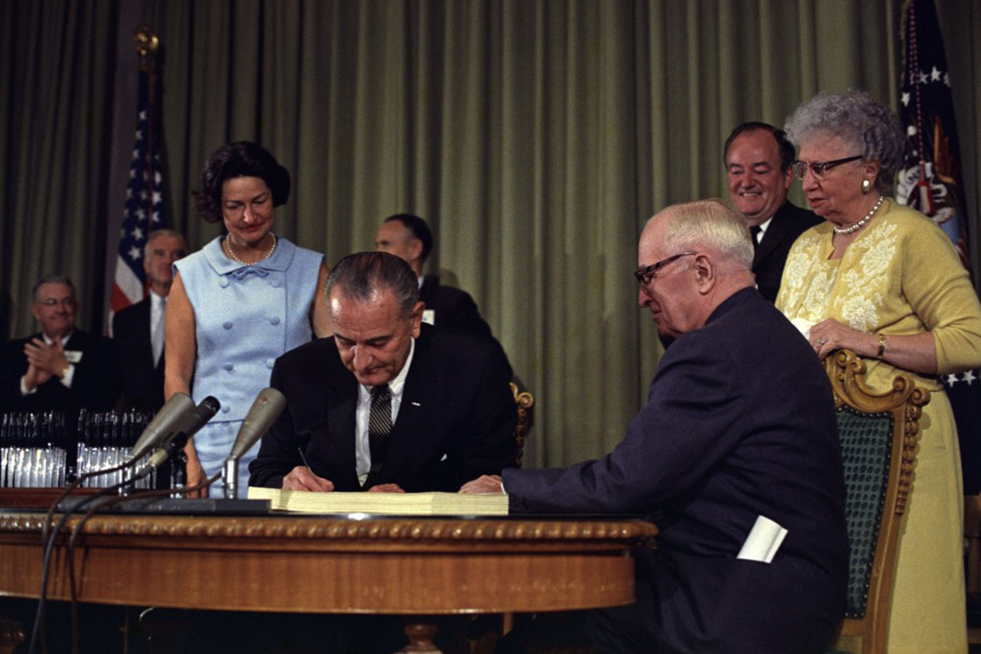 President Lyndon B. Johnson signing the Medicare Bill at the Harry S. Truman Library in Independence, Missouri. Former President Harry S. Truman is seated at the table with President Johnson. Lady Bird Johnson stands behind the president.