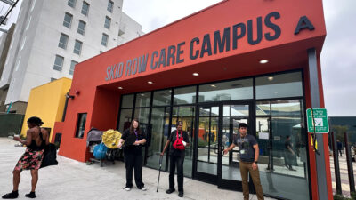 Four people standing in front of the Skid Row Care Campus in Los Angeles, a plave where people expriencing homelessness can get health care , find a place to rest, charge their phones, grab some food, or even get connected with housing.