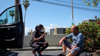 Street medicine practitioner speaks with man in a parking lot.